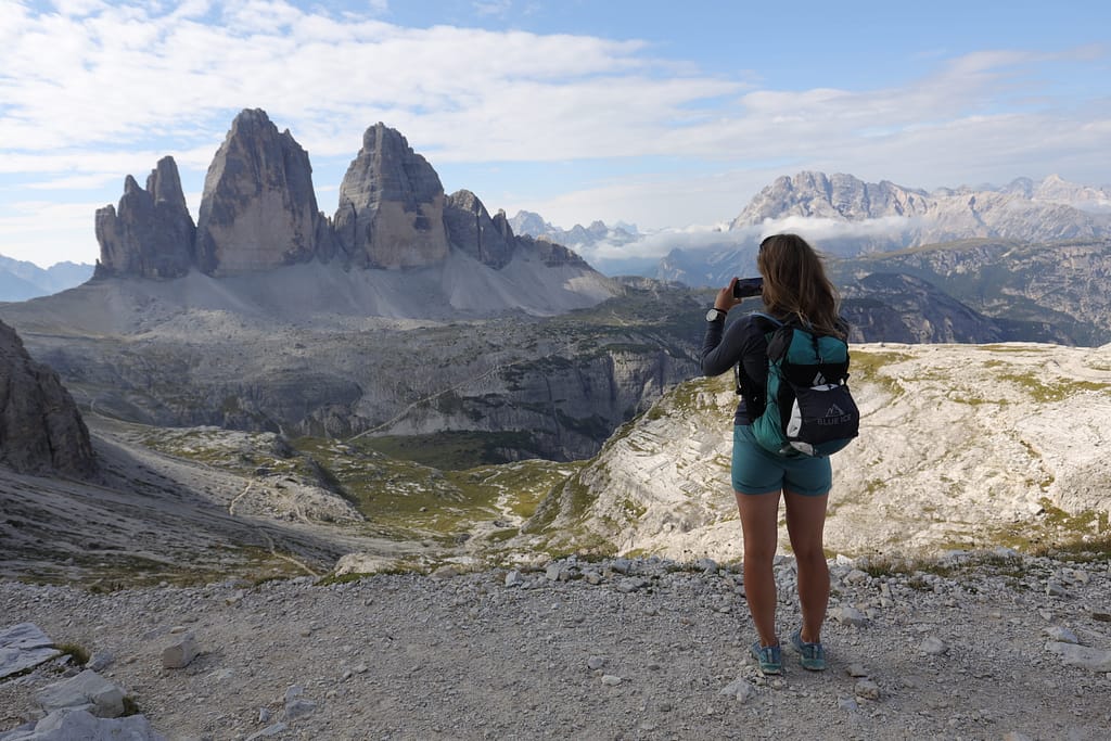 A woman in blue shorts and Black Diamond backpack taking picture of the iconic Tre Cime di Lavaredo towers