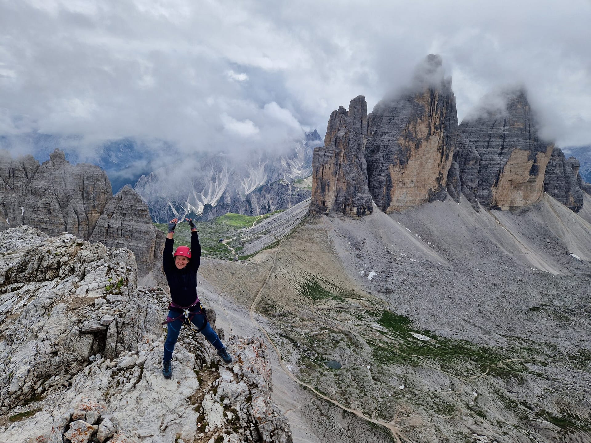 Woman with via ferrata gear standing on Monte Paterno summit with Tre Cime di Lavaredo towers emerging from the clouds behind her